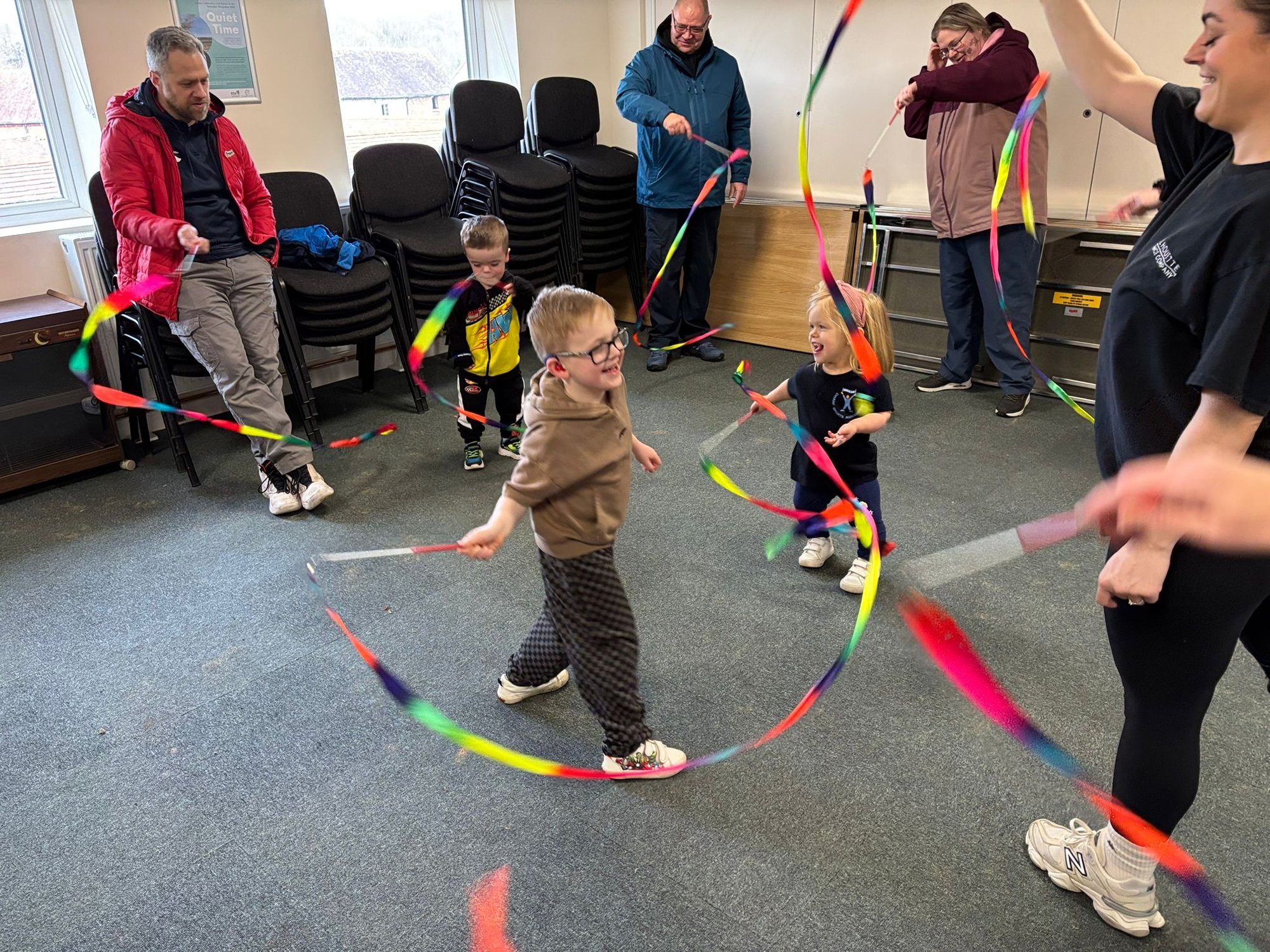 A photo of three Disabled children enjoying an activity with colourful ribbons inside a hall with their Parent Carers.