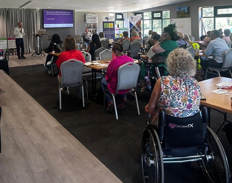 A photo taken at the back of the room towards the front, showing people attending the event watching a presentation taking place.