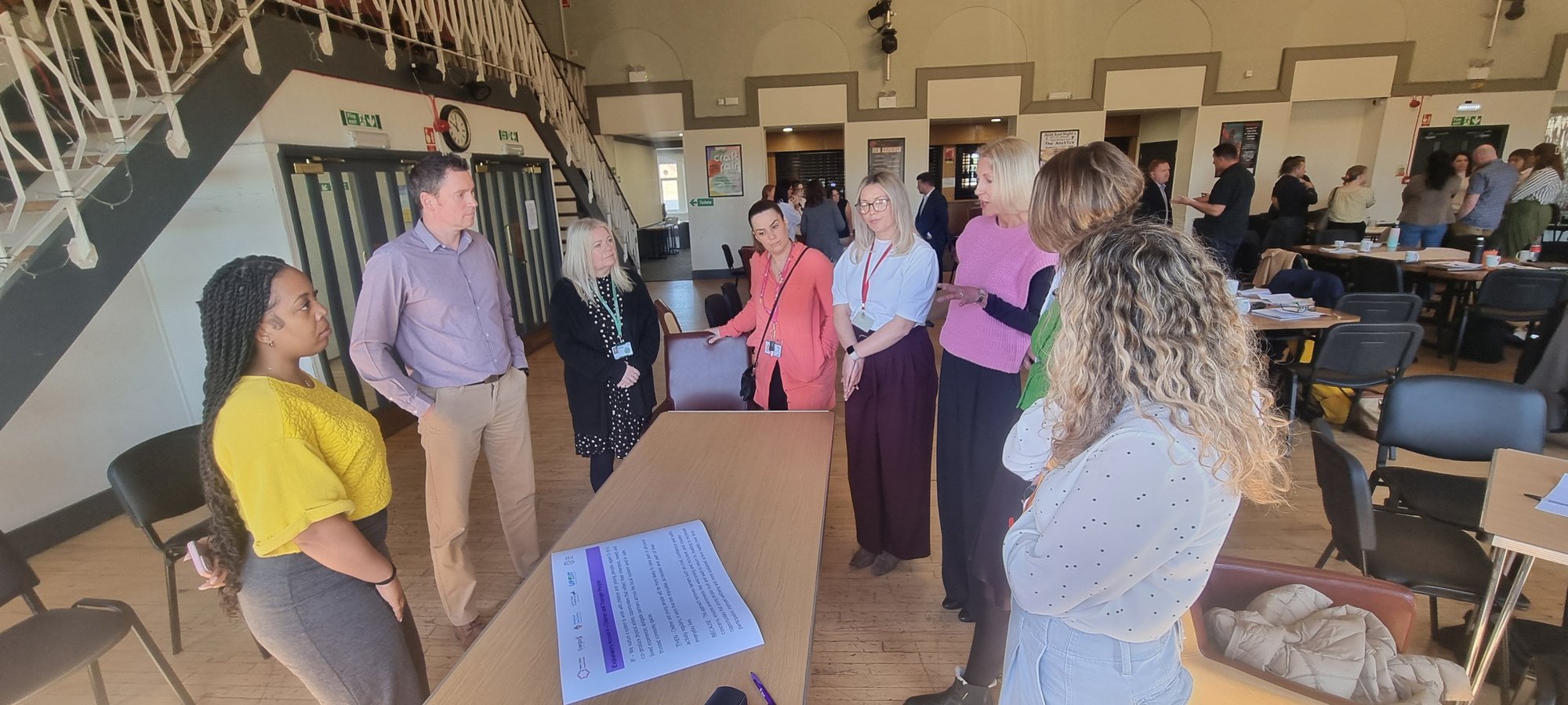 A group of 8 people (male and females) standing around a table in discussion as part of one of the System Leadership Training workshops.